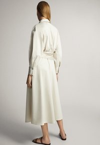 Robe longue beige avec une taille cintrée, des poignets boutonnés et un col. Texture lisse, atteignant le mi-mollet. Design simple et élégant.