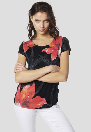 Juoda marškinėliai su dideliais raudonais gėlių raštais ir žaliomis detalėmis. Trumpomis rankovėmis ir apvalia iškirpte. Modelis pozuoja sukryžiavęs rankas.