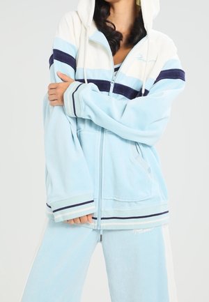 Frau trägt hellblauen und weißen Kapuzenpullover mit Reißverschluss und Marineblau gestreift, passende lockere Hose, verschränkte Arme, vor schlichtem hellem Hintergrund.