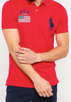 Mężczyzna w czerwonej koszulce polo z flagą USA i logo Ralph Lauren, ze skrzyżowanymi ramionami, na białym tle.