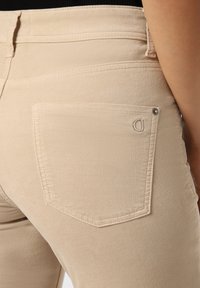Beige stretchjeans med en kroppsnära passform, bakficka med en subtil logodetalj och synliga sömnadsdetaljer. Mjuk textur.