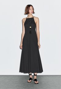 Sort maxi kjole med halterneck-design, i en flydende form, og en guldfarvet og sort kvastvedhæng. Sammen med sorte stroppsandaler.
