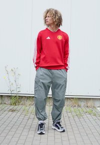 Rode sweatshirt met witte Adidas-strepen en logo, gecombineerd met losse grijze cargobroeken en zwart-witte sport sneakers.