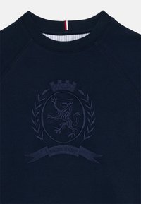Marineblå hettegenser med rund hals, med preget løveemblem, laurbærdetaljer og stripet kant ved halsen.