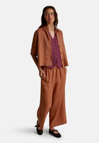 Donna che indossa pantaloni larghi color ruggine e una giacca a maniche corte sopra un gilet viola con bottoni, con scarpe piatte in rete nere e le mani in tasca.