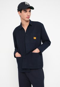 Granatowa koszulowa kurtka z bawełny z zapięciem na guziki, dwoma przednimi kieszeniami i małą pomarańczową metką na piersi. Długie rękawy i luźny krój.