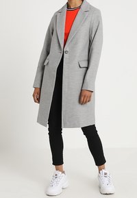 Harmaa pitkä takki, jossa on lovettu kaulus ja yksirivinen napitus, edessä kaksi taskua, päällä punainen ribbineulospusero ja mustat housut.