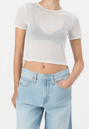 Femme portant un crop top blanc transparent à manches courtes par-dessus un soutien-gorge gris, assorti à un jean taille haute bleu clair, une main dans la poche.