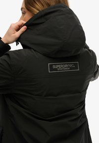 Veste noire imperméable avec capuche, arborant un logo brodé argenté "SUPERDRY NYC SPORTSWEAR" au dos et une finition texturée.