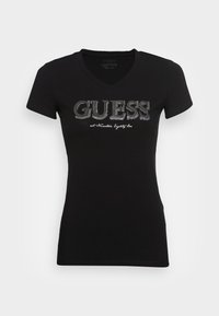 Schwarzes Baumwoll-T-Shirt mit V-Ausschnitt, das ein silbernes Strass-Logo "GUESS!" und weißen Text zeigt, kurze Ärmel und eine figurbetonte Passform hat.