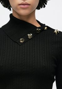 Schwarzes geripptes Strickoberteil mit hohem Kragen, verziert mit goldfarbenen dekorativen Knöpfen und einem strukturierten Zopfmuster.