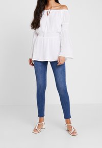 Blouse blanche à épaules dénudées avec un détail à nouer et une taille smockée, associée à un jean skinny bleu et des sandales blanches à lanières.