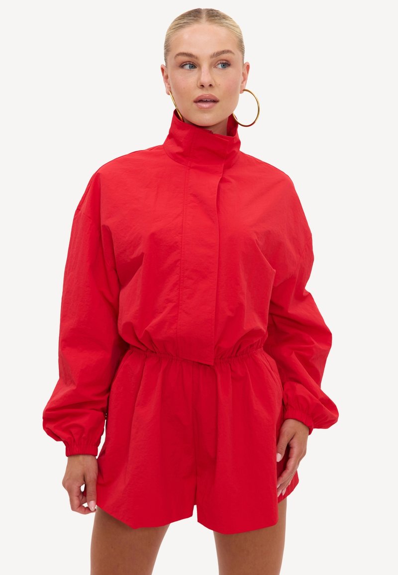 Femme aux cheveux blonds portant une combinaison rouge à manches longues avec un col haut, une taille élastique et de grandes boucles d'oreilles dorées, regardant sur le côté.