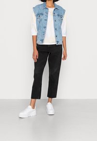 Kamizelka dżinsowa, biała koszula w wzory, czarne spodnie 3/4 i białe sneakersy. Kamizelka ma kieszenie i metalowe guziki; spodnie są proste.