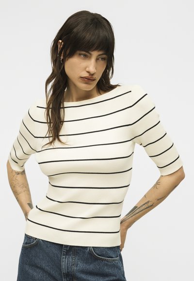 Γυναίκα με σκούρα μαλλιά, που φοράει άσπρο κοντομάνικο μπλουζάκι με μαύρες οριζόντιες ρίγες και μπλε τζιν, στέκεται με τα χέρια στη μέση.