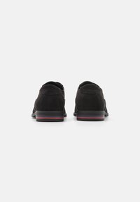Chaussures en daim noir avec un petit talon et un bout arrondi, présentant de subtiles rayures rouges et bleues sur le talon. Le design à l'arrière est lisse et épuré.