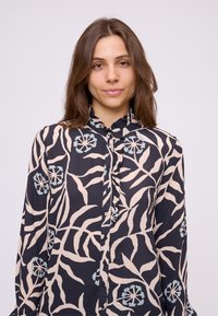 Giovane donna con lunghi capelli castani indossa una blusa blu navy con motivi floreali e foglie in rosa pallido e azzurro chiaro e colletto arricciato.