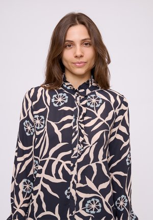 Giovane donna con lunghi capelli castani indossa una blusa blu navy con motivi floreali e foglie in rosa pallido e azzurro chiaro e colletto arricciato.