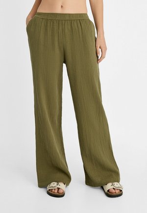 Pantalon ample vert olive texturé avec taille élastique porté par une personne les mains dans les poches, associé à des sandales beiges à boucle.