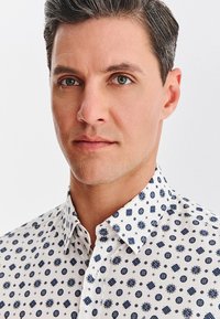 Biała koszula z niebieskimi, okrągłymi i kwiatowymi wzorami, z kołnierzykiem, zapinana na guziki z przodu, wykonana z gładkiej, lekkiej tkaniny.