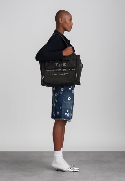 Borsa a tracolla nera in tela con "THE TOTE BAG" e "MARC JACOBS" stampati in grassetto. Presenta più scomparti e un'impugnatura robusta.