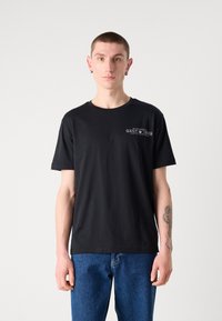 Black cotton t-shirt with short sleeves, crew neck, and a small graphic print on the left chest area. Paired with blue jeans.