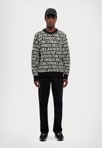 Maglione bianco e nero con design di testo ripetuto, abbinato a pantaloni neri e sneakers nere. Il modello indossa anche un cappellino nero.