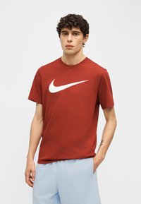 Νέος με σγουρά μαλλιά που φοράει κόκκινη μπλούζα Nike και ανοιχτό μπλε παντελόνι, στέκεται με το ένα χέρι στην τσέπη του μπροστά σε απλό φόντο.