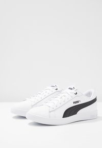 Para białych sneakersów Puma z czarnym paskiem bocznym i logo, zawiązanych, wystawionych na białej powierzchni i jasnoszarym tle.