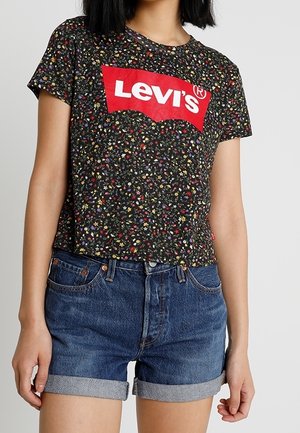 Kobieta w czarnym kwiatowym T-shircie Levi’s z czerwonym logo i niebieskich podwiniętych szortach jeansowych na jednolitym tle.