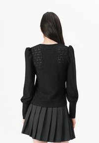 Pullover in pizzo nero a maglia con maniche a palloncino lunghe, caratterizzato da un motivo a diamante. Indossato con una gonna grigia plissettata.