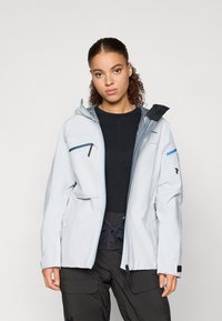 Nuori nainen, jolla on kihara hius ja päällään vaaleanharmaa ulkoilutakki mustan paidan ja mustien cargo-housujen kanssa, seisoo yksinkertaista taustaa vasten.