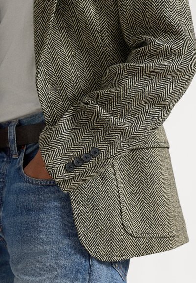 Rękaw marynarki w jodełkę z czterema czarnymi guzikami, noszony na wpuszczonej szarej koszuli i niebieskich dżinsach z czarnym paskiem.