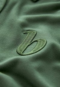 Tessuto verde con una superficie liscia caratterizzato da una lettera "b" ricamata in una tonalità corrispondente. Il design enfatizza una sottile eleganza e semplicità.