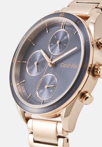 Montre en acier inoxidable doré avec un cadran texturé bleu, trois sous-cadrans et un bracelet en métal assorti. Nom de la marque "Calvin Klein" en relief.