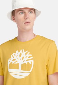 Žluté bavlněné tričko s velkým bílým logem stromu na přední straně. Design je jednoduchý, s klasickým crew výstřihem a krátkými rukávy.