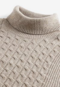 Beige polo-tröja med en ribbad krage och en texturerad flätstickad mönster, tillverkad av ett mjukt, varmt material med en subtil glans.