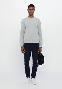 Sudadera gris con logo en relieve, pantalones de chándal azul marino con rayas laterales, zapatillas blancas y una pequeña bolsa negra en la mano.