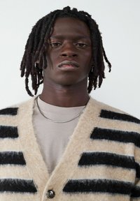 Beżowy kardigan w czarne paski, wykonany z miękkiego materiału, z zapięciem na guziki i dekoltem w serek. Nosi się go na jasnoszarej koszuli.