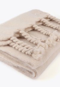 Beige tæppe lavet af blødt materiale, med teksturerede fletninger og pom-pom detaljer langs én kant, pænt foldet til udstilling.