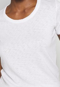 Camiseta blanca de tela suave y texturizada con un escote amplio y redondeado, que presenta mangas cortas y un ajuste relajado.