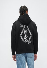 Un hoodie noir en mélange de coton qui présente un grand graphique blanc au dos, représentant un design de main et de chaîne. Poignets et ourlet côtelés.