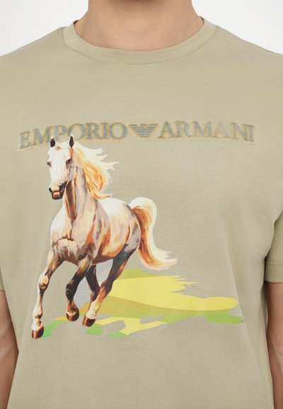 T-shirt en coton beige avec un imprimé coloré d'un cheval en course et le logo "EMPORIO ARMANI" en lettres dorées au-dessus du graphique.