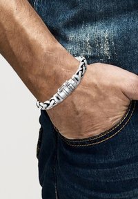 Metall- og læderflettet armbånd med en sølvfarvet lås, i en sort og hvid farveskema, sat mod denimstof.