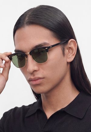 Persona con i capelli lunghi e scuri indossa occhiali da sole neri e dorati e una camicia nera con colletto, aggiustando gli occhiali con una mano.