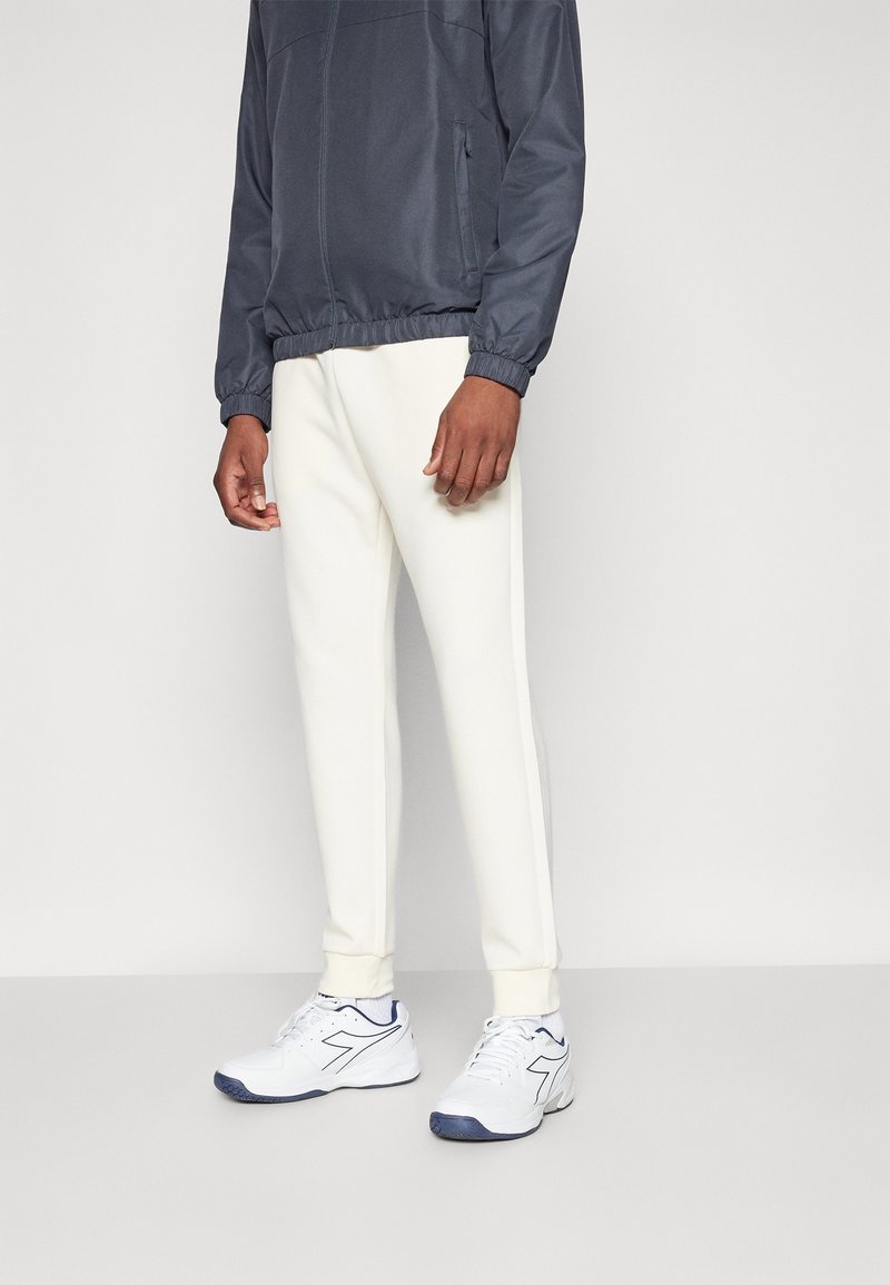 Mansportoutfit bestående av en mörkgrå jacka, vita fleecebyxor och vita sneakers med marinblå detaljer. Mjuk textur, avslappnad design.