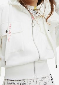 Felpa bianca con zip frontale, due tasche anteriori e coulisse. Presenta un dettaglio di testo ricamato rosso sul lato sinistro e orlo a costine.