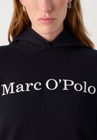 Schwarzer Kapuzenpullover aus weichem Material mit einem großen weißen Logo "Marc O'Polo", das prominent auf der Vorderseite aufgedruckt ist.