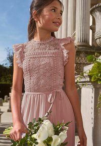 Világos rózsaszín ruha csipkebetétes, virágmintás felsőrésszel és fodros ujjakkal, derékban megkötve, és légies szoknyával. Fehér rózsákból álló csokrot tart.