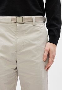 Ανοιχτό μπεζ παντελόνι με υφή, που διαθέτει μεταλλική πόρπη ζώνης και επίπεδη ζώνη, παρουσιαζόμενο με μαύρο πουκάμισο μακρυμάνικο.